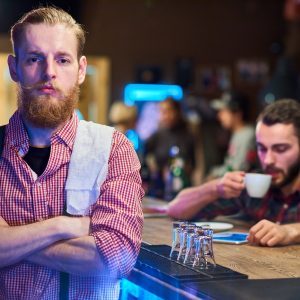 Portrait of handsome bearded bartender posing with arms crossed looking confidently at camera standing behind bar counter in pub, copy space