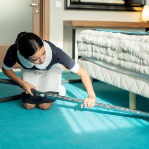 Young beautiful brunette chambermaid in uniform using vacuum cleaner while cleaning blue floor covering under bed in hotel room