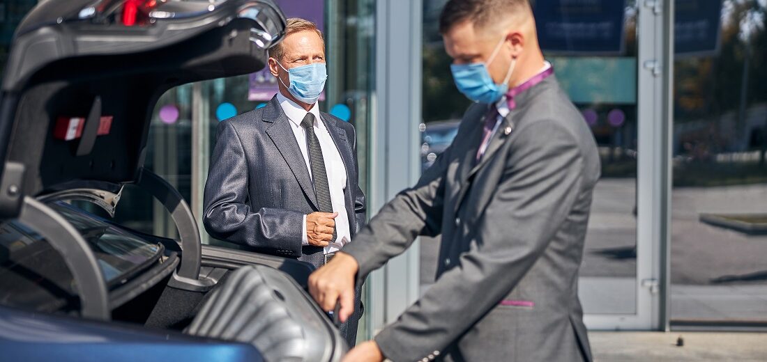 Young man in suit and mask packing businessman luggage into car trunk after his arrival to airport