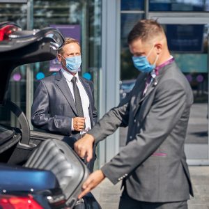 Young man in suit and mask packing businessman luggage into car trunk after his arrival to airport